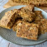 thermomix anzac slice on grey plate, tray of slice in background. on white wood