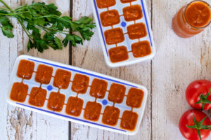 Thermomix tomato paste in ice cube trays and jar on white wood with parsley and tomatoes on vine