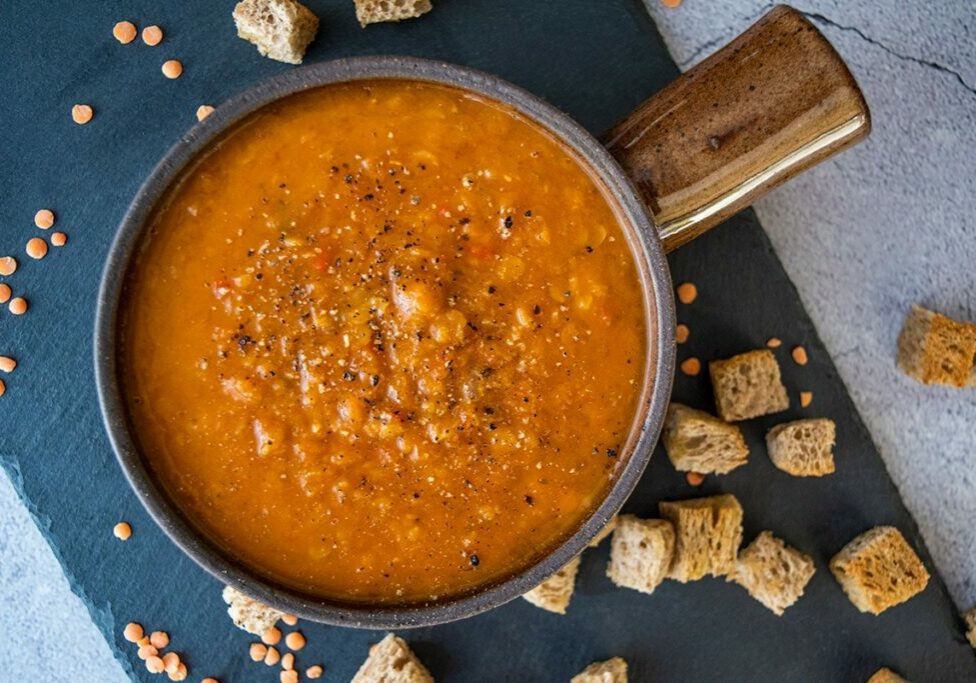 spicy tomato and lentil soup made in thermomix, in brown bowl with handle. Sitting on black slate tile on grey backdrop, with croutons and dry lentils in on the tile